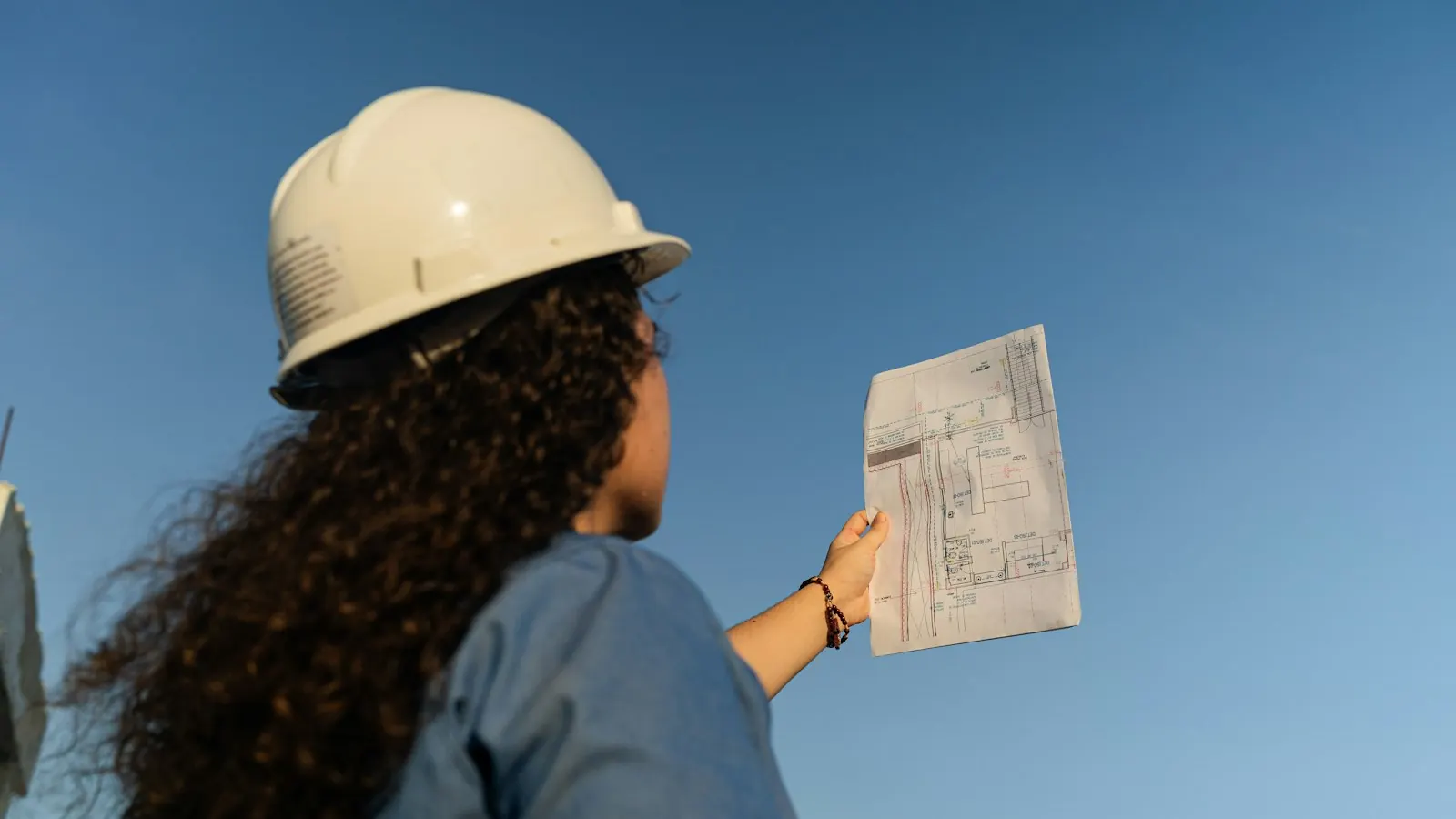A construction professional in a hard hat holding up building plans against a blue sky